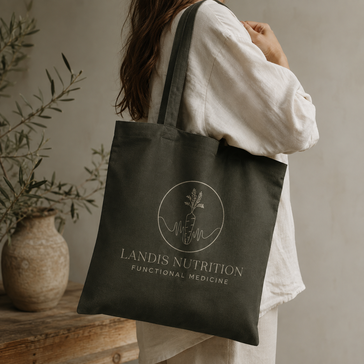 Editorial lifestyle mockup of a woman in a cream sweater carrying a dark olive canvas tote featuring the Landis Nutrition & Functional Medicine logo in a warm, earthy setting with natural greenery and textured stone.